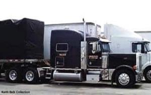 Black colored flat bed truck is standing with different type of freight truck